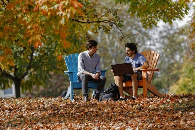 two students sitting in colorful Adirondack chairs with fall colored leaves surrounding them 