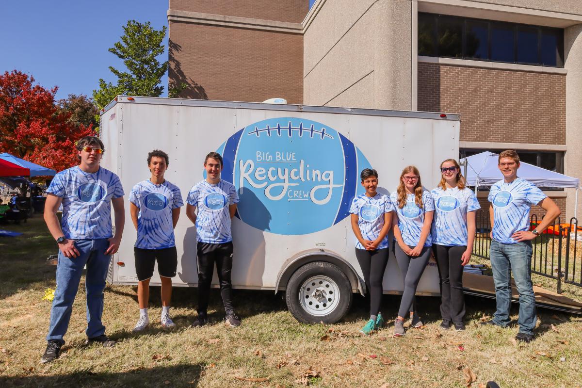 seven people standing outside in front of a trailer that has a football icon with the words Big Blue Recycling
