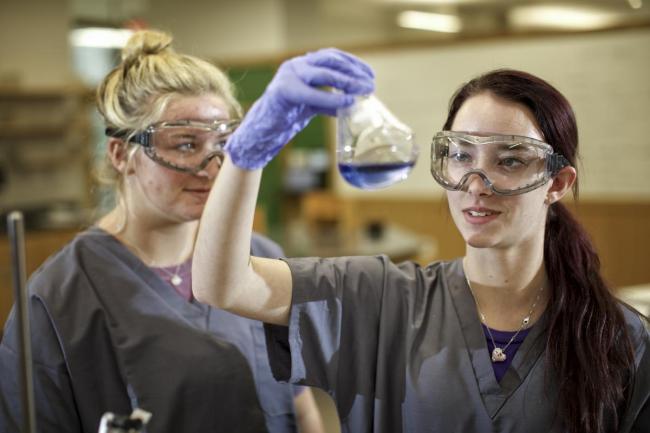 Two female students in a lab looking at a beaker filled with a purple liquid