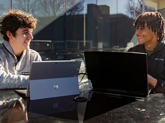 Two male students sitting at a table outside looking at a computer