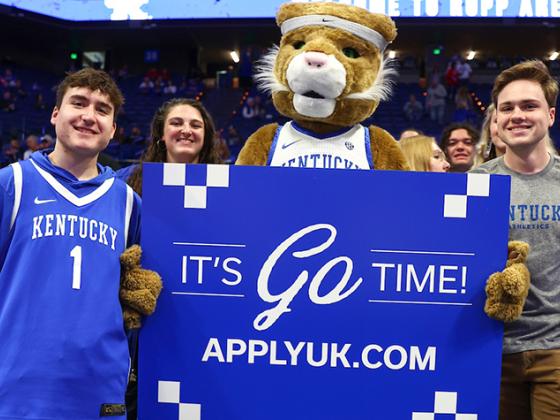 Wildcat Mascot in the student section at basketball holding a sign that says apply